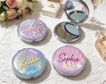 May include: Four personalized compact mirrors with glitter and names: Theresa, Kristin, and Sophia. One mirror is open, revealing a reflective surface. The mirrors are silver-toned with a shimmering, colorful design. A ring box, flowers, and a glass of champagne are in the background.
