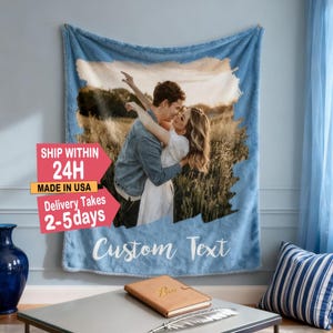 Op de afbeelding: Een lichtblauwe deken met een foto van een stel dat elkaar omhelst in een veld. De deken heeft een lichtblauwe rand en de woorden "Custom Text" in witte letters. De deken hangt aan een muur.
