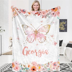 May include: A white blanket with a pink butterfly design and the name "Georgia" in pink script. The blanket features a floral border and accents, with gold details on the butterfly.