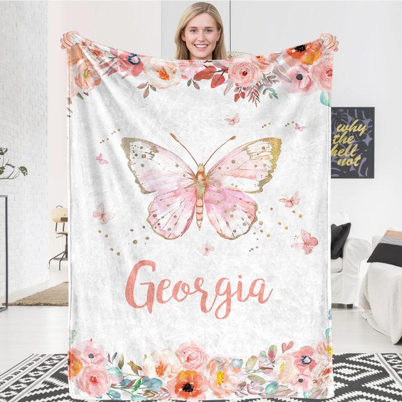 May include: A white blanket with a pink butterfly design and the name "Georgia" in pink script. The blanket features a floral border and accents, with gold details on the butterfly.