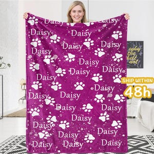 May include: A pink fleece blanket with a white paw print pattern and the word "Daisy" repeated throughout. The blanket is soft and cozy, perfect for cuddling up on the couch.