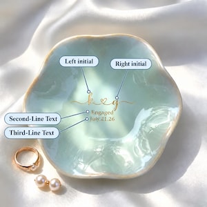 May include: A light blue, scalloped ceramic ring dish featuring gold script text that reads "k & g", "Engaged", and "July.21.26". A gold ring with a white pearl and a pair of matching pearl earrings are placed beside the dish.