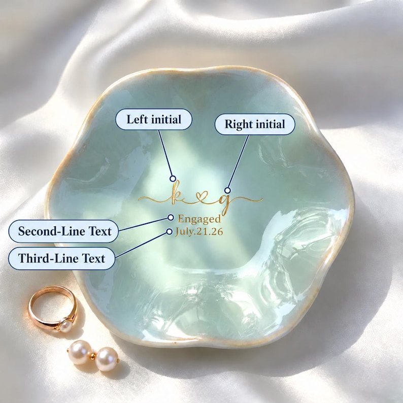 May include: A light blue, scalloped ceramic ring dish featuring gold script text that reads "k & g", "Engaged", and "July.21.26". A gold ring with a white pearl and a pair of matching pearl earrings are placed beside the dish.