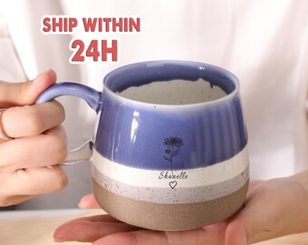 May include: A handmade ceramic mug with a blue, white, and gray color scheme. The mug features a floral design and the name "Sharelle" with a heart symbol. The mug has a handle and a brown base.