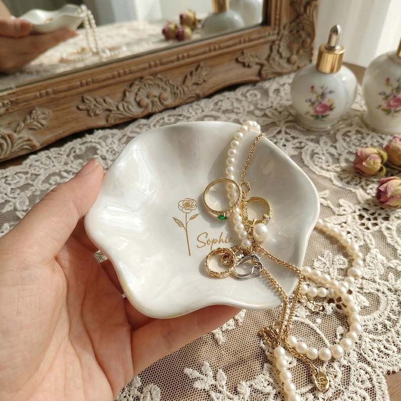 May include: A white, scalloped ceramic trinket dish featuring a gold-colored line drawing of a rose and the name Sophia printed in gold script. The dish contains several gold-toned rings and a long strand of white pearls draped over the edges.