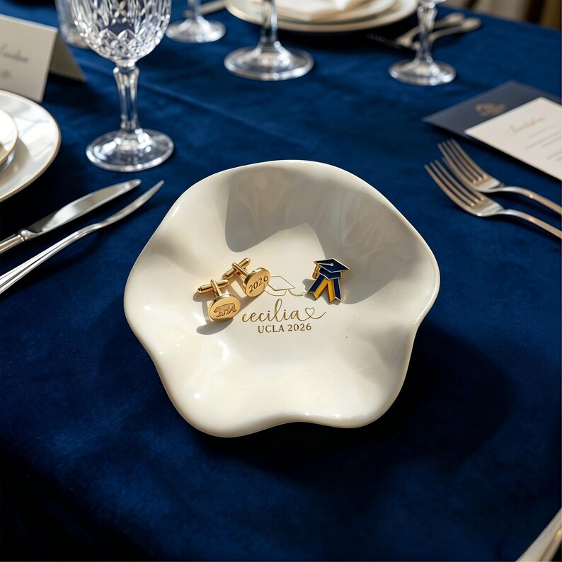 Puede incluir: Un plato de cer&aacute;mica blanco con borde festoneado con gemelos dorados y un pin de graduaci&oacute;n. El plato est&aacute; inscrito con "Cecilia UCLA 2026". Los gemelos y el pin son dorados con detalles azules y amarillos.