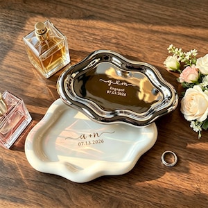 May include: Two decorative trays with wavy edges, one silver and one white, with engraved text and dates. A ring and two perfume bottles are also present. The silver tray reads "g & m Engaged 07.03.2026". The white tray reads "a + n 07.13.2026".