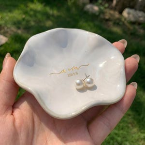 May include: A white, scalloped-edge ceramic trinket dish featuring gold cursive text that reads a + m and the date 2026.5.18. A pair of white pearl stud earrings with silver posts rests inside the center of the glossy, shell-shaped dish.