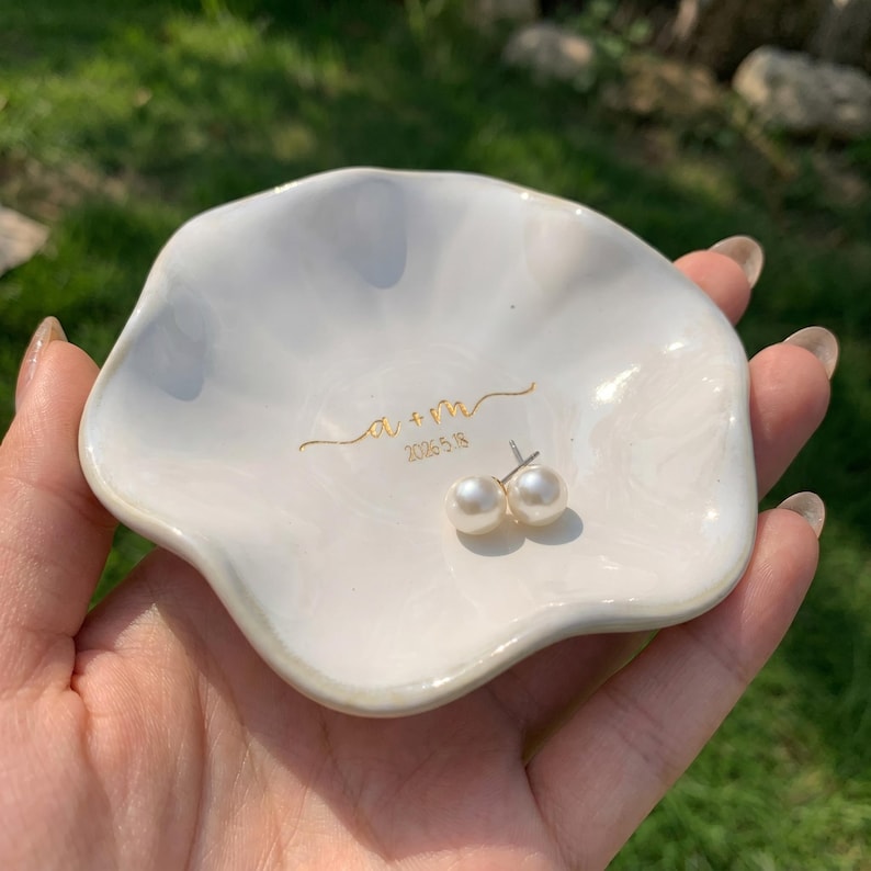 May include: A white, scalloped-edge ceramic trinket dish featuring gold cursive text that reads a + m and the date 2026.5.18. A pair of white pearl stud earrings with silver posts rests inside the center of the glossy, shell-shaped dish.