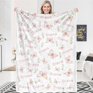 May include: A white blanket featuring a repeating pattern of pink butterflies, floral accents, and the name "Diana" in a pink script. The blanket is held up by a person.