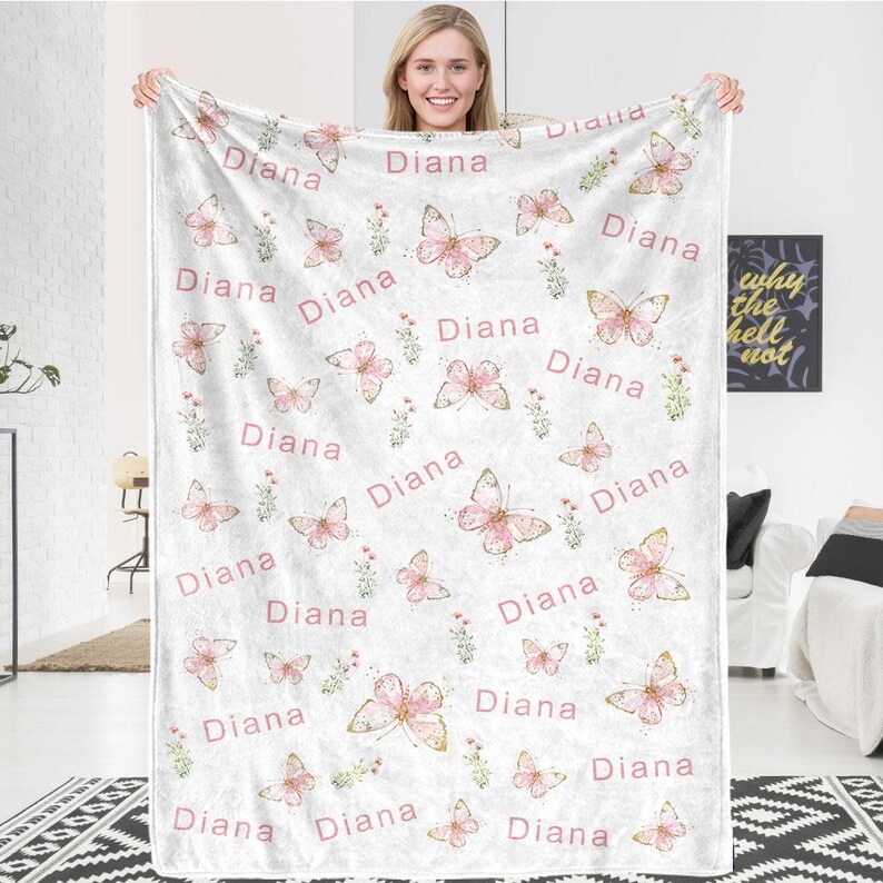 May include: A white blanket featuring a repeating pattern of pink butterflies, floral accents, and the name "Diana" in a pink script. The blanket is held up by a person.