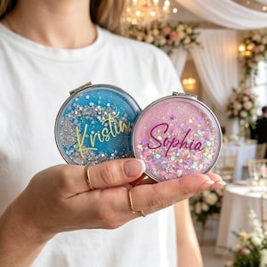 Puede incluir: Dos espejos compactos personalizados, uno azul con "Kristin" y otro rosa con "Sophia" en cursiva. Ambos espejos tienen purpurina plateada y detalles de estrellas. Los compactos se sostienen en una mano, con un fondo borroso.