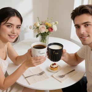 May include: A white mug with the name "Sophia" and a black mug with the name "Kate" are held by two people. The white mug contains dark liquid, and the black mug contains a white liquid. A small cupcake and flowers are on the table.