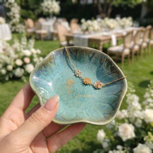 May include: A small, scalloped ceramic trinket dish with a glossy teal and blue glaze. The dish features a gold-colored line drawing of a sunflower and the name Cecilia in script. A delicate gold-tone chain bracelet with flower charms rests inside.