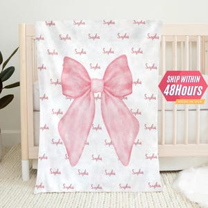 May include: A white baby blanket with a pink watercolor bow in the center. The blanket is covered in the name "Sophia" repeated in a pink script font.