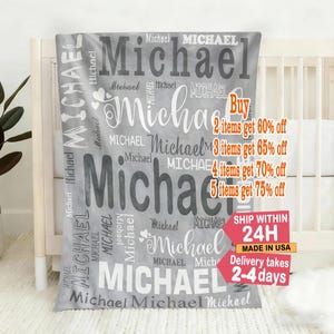 May include: A grey blanket featuring the name "Michael" in different fonts and sizes. The blanket is draped over a cot. The name "Michael" is printed in both dark and light grey. The blanket appears soft.