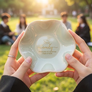 Puede incluir: Un peque&ntilde;o plato de cer&aacute;mica blanquecina con borde festoneado. El plato presenta texto dorado que dice "emily" con un icono de gorro de graduaci&oacute;n, y "University of California Class of 2026".