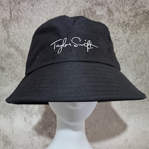 May include: Black bucket hat with the white embroidered signature "Taylor Swift".