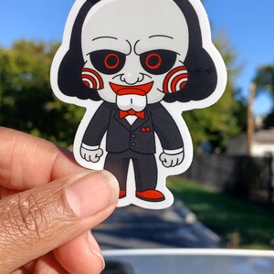 May include: A sticker featuring a cartoon character with a white face, black hair, and red eyes. The character wears a black suit with a red bow tie and red shoes. The sticker is held up against a blue sky.