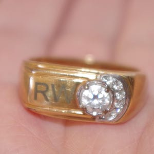 Könnte beinhalten: Ein goldener Ring mit einem großen, klaren, runden Diamanten und kleineren Akzentdiamanten. Der Ring hat die Buchstaben "RW" auf der Seite eingraviert. Der Ring wird in einer Hand gehalten.