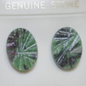 Może przedstawiać: Dwa owalne kaboszony z prawdziwego kamienia. Kamienie są zielone z ciemnozielonymi i fioletowymi wtrąceniami oraz rzeźbionym wzorem. Tekst "GENUINE STONE" znajduje się nad kamieniami.
