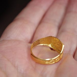 May include: A gold ring with a rectangular face and intricate detailing. The ring is held in an open hand, showcasing its bright, polished finish. The band features a textured design.