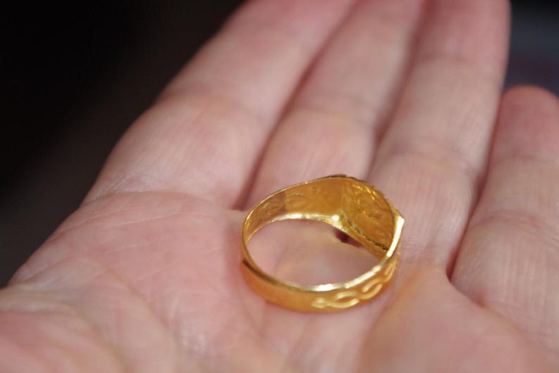 May include: A gold ring with a rectangular face and intricate detailing. The ring is held in an open hand, showcasing its bright, polished finish. The band features a textured design.