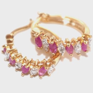 May include: A pair of gold hoop earrings, each adorned with alternating marquise-cut ruby and diamond gemstones. The earrings are set in gold and are presented against a white backdrop. The gemstones are pink and clear.