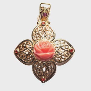 May include: A gold pendant featuring a central carved pink coral rose surrounded by four heart-shaped filigree sections. The piece is accented with small red gemstones set into the filigree and the bail, which has a polished gold finish.