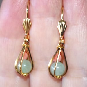 May include: A pair of gold-colored drop earrings. Each earring features a teardrop-shaped cage design, holding a light green and a coral-colored bead. The earrings have a decorative shell-like element at the top.
