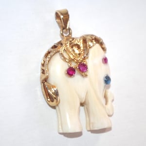 May include: Ivory-coloured elephant pendant with gold accents and gemstone details. The pendant features a gold bail, ornate gold detailing on the head, and ruby and sapphire gemstones. The elephant has a trunk and tusks.