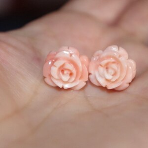 Puede incluir: Dos delicados pendientes en forma de rosa rosa claro. Los pendientes est&aacute;n tallados con detalles intrincados, mostrando los p&eacute;talos de las rosas. Los pendientes est&aacute;n colocados sobre un fondo borroso, resaltando el dise&ntilde;o de la joya. Estos pendientes son un hermoso accesorio.