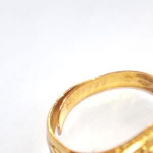 May include: Close-up of a gold ring with engraved details. The ring's interior displays markings, including the number "9999". The ring has a rectangular face with a detailed design. The ring is set against a white background.