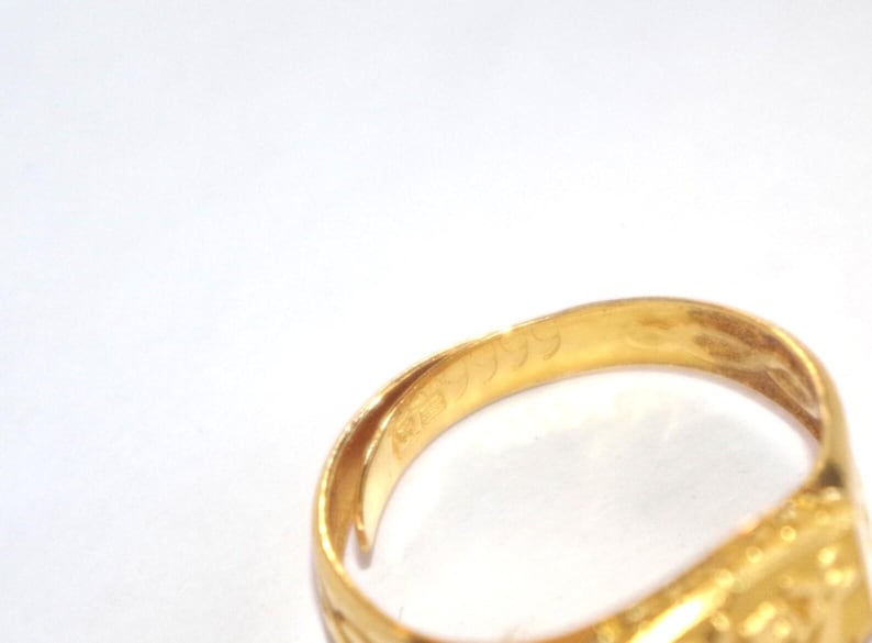 May include: Close-up of a gold ring with engraved details. The ring's interior displays markings, including the number "9999". The ring has a rectangular face with a detailed design. The ring is set against a white background.