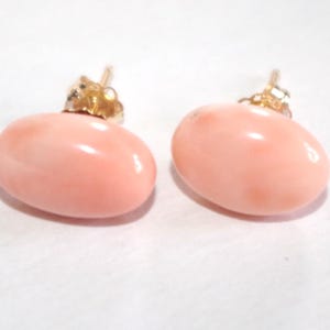 Puede incluir: Un par de pendientes ovalados de color rosa pálido con postes dorados. Los pendientes tienen una superficie lisa y pulida y un ligero brillo. Las joyas están sobre un fondo blanco.