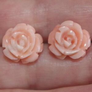 Puede incluir: Dos pendientes en forma de rosa de color rosa claro. Los pendientes est&aacute;n tallados con p&eacute;talos detallados, mostrando un delicado dise&ntilde;o floral. El color es un rosa suave y natural, y los pendientes parecen estar hechos de un material liso.