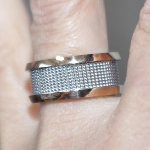 May include: A wide ring with a silver mesh center band and gold-colored edges. The ring is shown on a finger, highlighting the contrast between the metallic elements and the texture of the mesh.