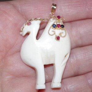 May include: Ivory-colored camel pendant with gold accents and gemstone embellishments. The pendant features a detailed camel design with a gold chain and a decorative gold element adorned with red and blue gemstones. A small pink gemstone is on the camel's head.