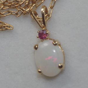 May include: A gold-colored necklace with a pendant featuring a large, oval, white opal gemstone. A small, round, pink gemstone sits above the opal. The necklace chain is also gold-colored.