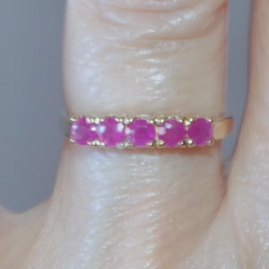 Puede incluir: Un anillo de oro con seis piedras preciosas rosas brillantes de talla ovalada. Las piedras están engastadas juntas, creando una banda continua de color. El anillo se muestra en el dedo de una persona.