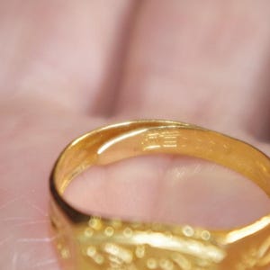 May include: Close-up of a gold ring. The ring is a bright, shiny gold color. The ring has a smooth, polished surface with some etched details. The ring is held in a person's hand.