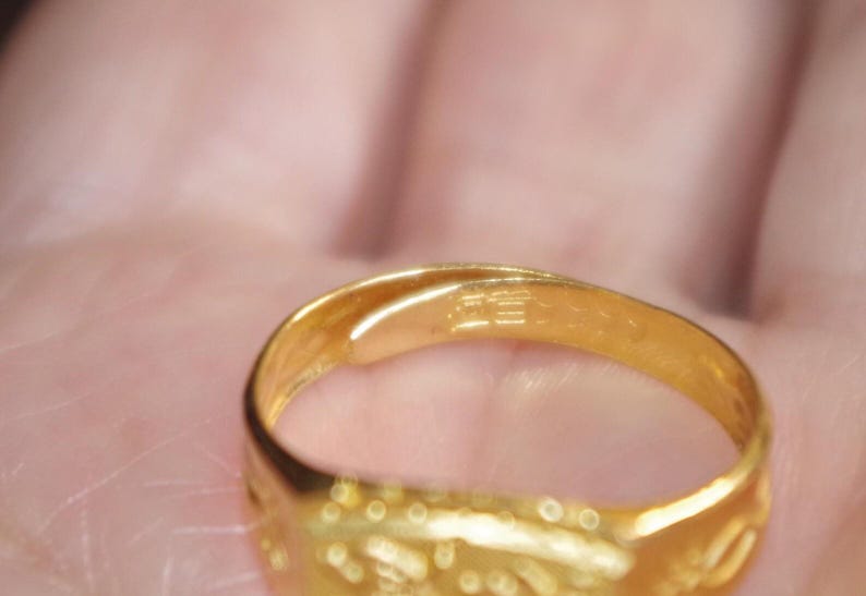 May include: Close-up of a gold ring. The ring is a bright, shiny gold color. The ring has a smooth, polished surface with some etched details. The ring is held in a person's hand.