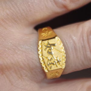 May include: A gold ring with an engraved design. The ring has an oblong face with a textured border and decorative elements. The ring is a bright gold color and appears to be worn on a finger.