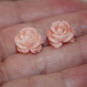 Puede incluir: Dos peque&ntilde;os objetos tallados en forma de rosa, de color melocot&oacute;n claro. Las tallas detalladas muestran los p&eacute;talos de las rosas. Los objetos se sostienen en una mano, lo que sugiere su peque&ntilde;o tama&ntilde;o. Las rosas parecen estar hechas de piedra u otro material duro.