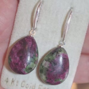 May include: A pair of teardrop earrings with silver hooks. The earrings have polished stones in deep red, green, and grey. They are displayed on a light green card. The earrings are a unique accessory.