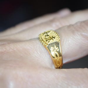 May include: A gold ring with Chinese characters and floral designs. The ring is on a finger, showcasing its intricate details. The ring's rectangular top features Chinese characters, and the band has a floral pattern.