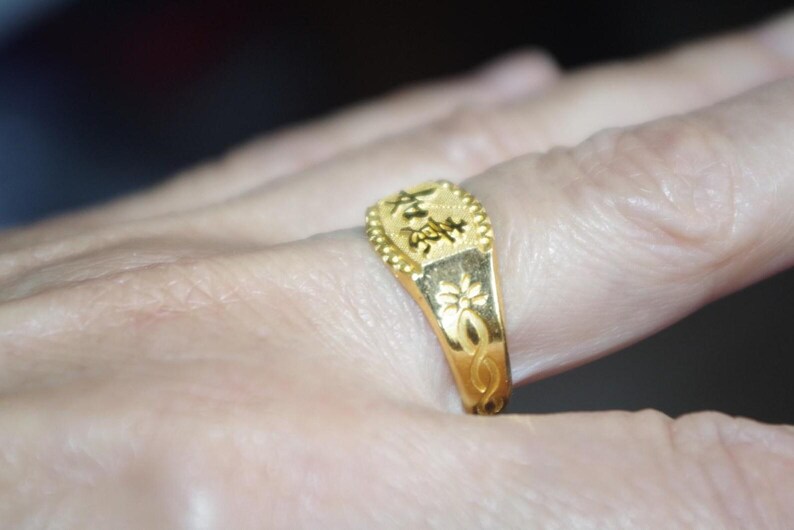 May include: A gold ring with Chinese characters and floral designs. The ring is on a finger, showcasing its intricate details. The ring's rectangular top features Chinese characters, and the band has a floral pattern.