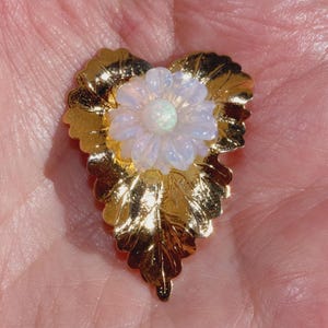 May include: A gold-toned brooch shaped like a heart with a textured leaf pattern. Centered on the heart is a white, translucent, multi-petaled flower with a round, iridescent cabochon at its core, reflecting subtle blue and green light tones.