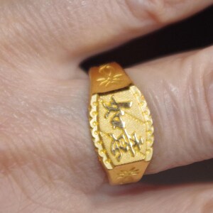 May include: A gold-colored ring with an engraved design. The ring has an oblong face with a textured border and decorative elements. The ring is worn on a finger.