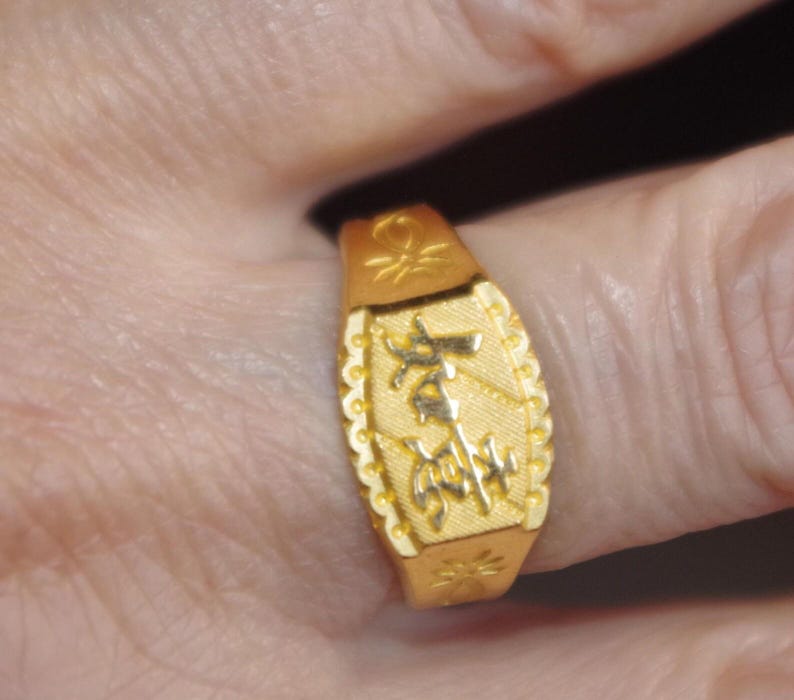 May include: A gold-colored ring with an engraved design. The ring has an oblong face with a textured border and decorative elements. The ring is worn on a finger.